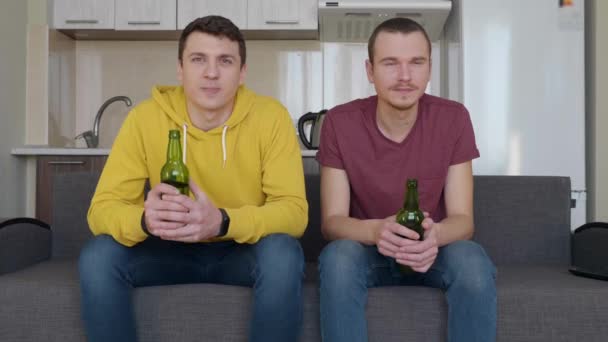 Dos hombres sentados en el sofá y viendo un partido de fútbol en la televisión y sosteniendo botellas de cerveza en sus manos. Dos jóvenes mirando tranquilamente un partido de deportes en el sofá. Una cocina en el fondo. Imágenes 4K
. - Metraje, vídeo
