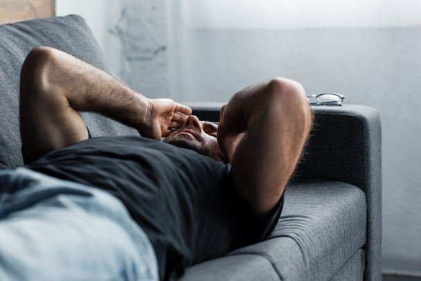 depressed man lying on grey sofa with closed eyes and hands near head - Photo, Image