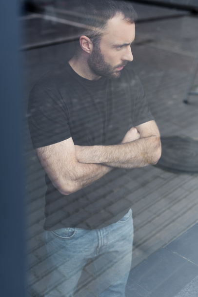 όμορφος σοβαρός άνθρωπος με σταυρωτά χέρια στέκεται στο παράθυρο και κοιτάζει μακριά  - Φωτογραφία, εικόνα