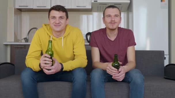 Dva muži sedí na gauči a sledují fotbalový zápas v televizi, pijí pivo, komentují a usmívají se. Dva mladí chlápci klidně sledují sportovní zápas na gauči. Kuchyň na backgrogu. 4k záběr - Záběry, video