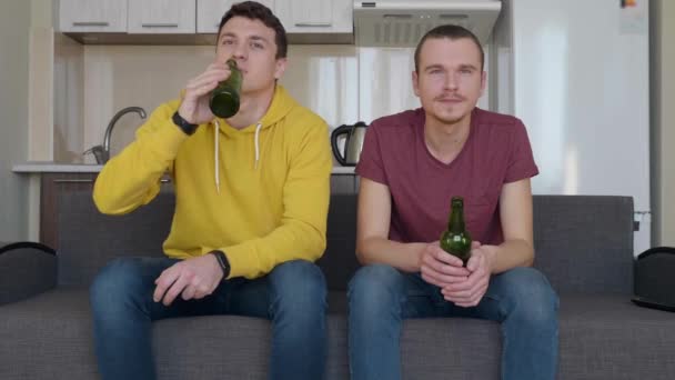 Zwei Männer sitzen auf der Couch und schauen sich ein Fußballspiel im Fernsehen an und trinken Bier. zwei junge Kerle, die gelassen ein Sportspiel auf dem Sofa verfolgen. eine Küche im Hintergrund. 4k Filmmaterial. - Filmmaterial, Video