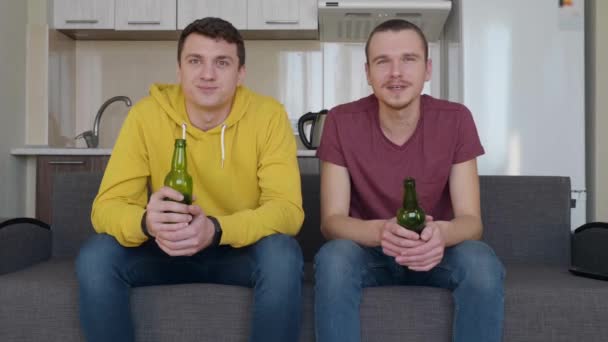 Two men siting on the couch and watch a football match on TV, drink beer, commenting and smile. Two young cheerful guys calmly watching a sports game and talking. Kitchen on the backgroung. 4K footage - Footage, Video