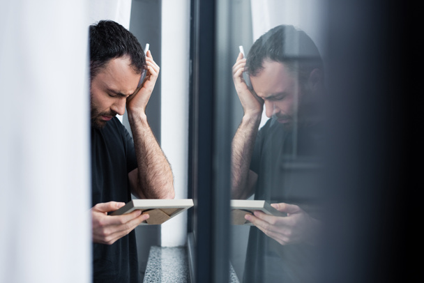 selectieve focus van huilen man vasthouden fotolijstjes terwijl staande door venster - Foto, afbeelding