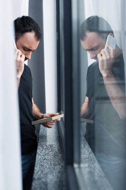 selective focus of upset handsome man with smartphone holding photo frame while standing by window - Photo, Image