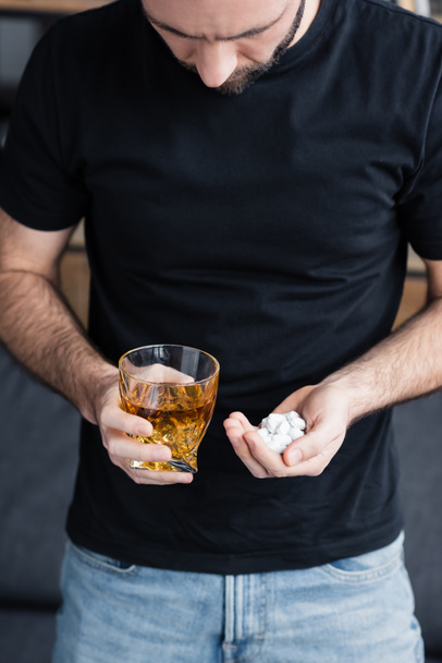 cropped view of depressed man in black t-shirt with handful and pills and glass of whiskey - Photo, Image