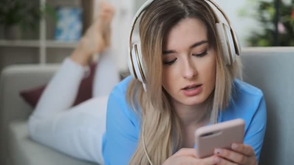 Young beautiful girl in blue outfit enjoying the music at home. Woman listening music in headphones from smartphone and dancing while lying on sofa in room. - Video, Çekim