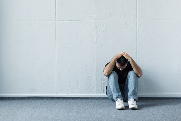 homem deprimido sentado no chão perto da parede branca e de mãos dadas na cabeça
 - Foto, Imagem