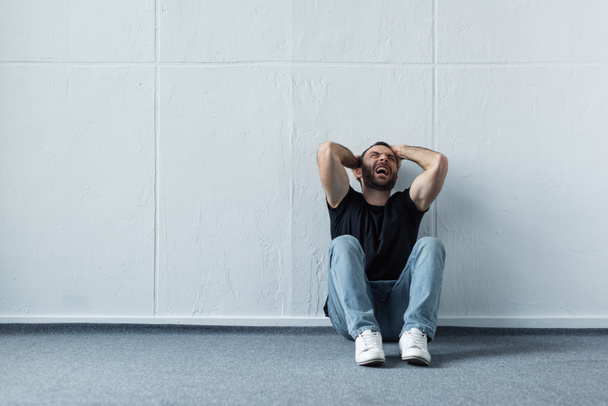 adulto deprimido hombre gritando mientras sentado en el suelo por la pared blanca
  - Foto, Imagen