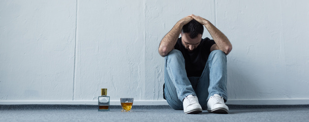 panoramische shot van lijdende man zittend op de vloer door witte muur in de buurt van fles en glas whisky - Foto, afbeelding