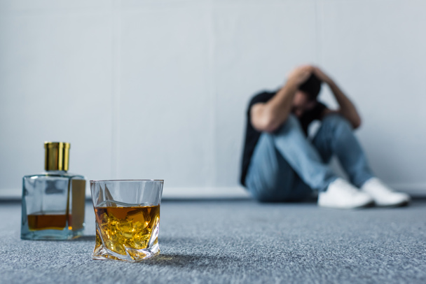 enfoque selectivo del hombre que sufre sentado en el suelo por la pared blanca cerca de la botella y el vidrio con whisky
 - Foto, Imagen