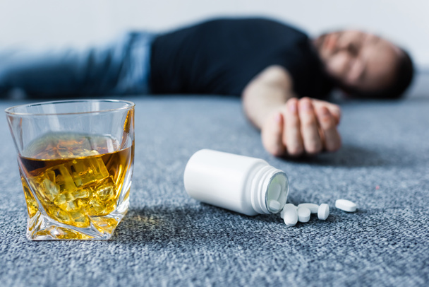 enfoque selectivo del hombre inconsciente acostado en el suelo cerca del vaso de whisky y recipiente con pastillas
  - Foto, imagen