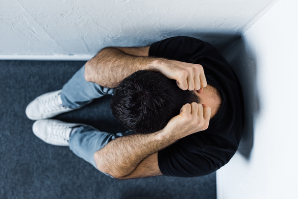 top view of adult depressed man sitting on floor in corner and holding hands on head - Fotó, kép