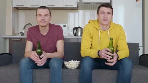 Dos hombres sentados en el sofá ven un partido de fútbol en la televisión, beben cerveza y comen palomitas. Dos jóvenes mirando tranquilamente un partido de deportes. Una cocina en el fondo. Imágenes 4K
 - Metraje, vídeo