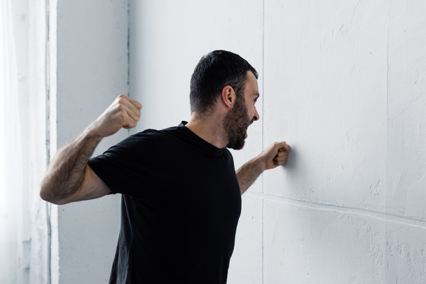 angry bearded man in black t-shirt screaming and kicking white wall - Фото, изображение