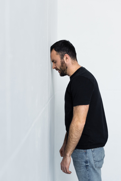 sfrustrowany Brodaty człowieka w czarnym t-shirt stojący w pobliżu białej ścianie z zamkniętymi oczami - Zdjęcie, obraz