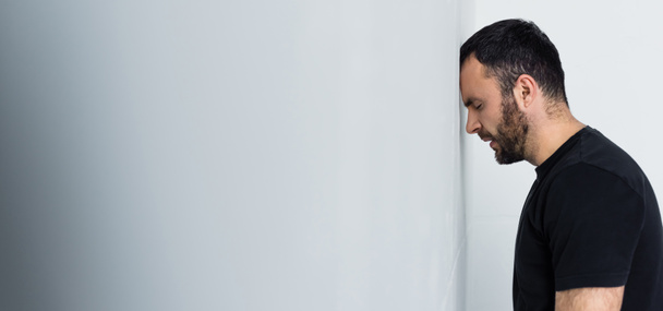 tiro panorâmico de homem barbudo deprimido em pé junto à parede branca com os olhos fechados
 - Foto, Imagem