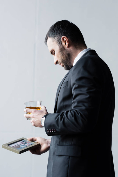 upset bearded man in suit holding photo in frame and glass of whiskey - Fotoğraf, Görsel