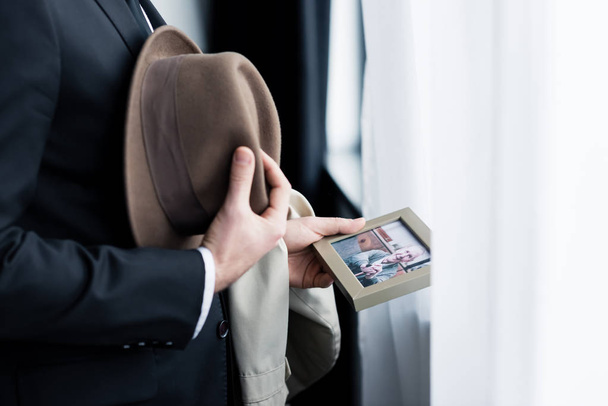 cropped view of man holding photo in frame while standing by window with hat in hand - 写真・画像