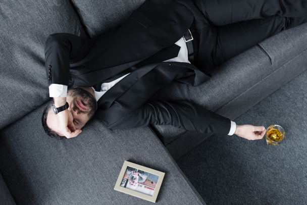 Top uitzicht van gefrustreerde zakenman liggend op de Bank in de buurt van foto in frame en glas whisky op de vloer - Foto, afbeelding