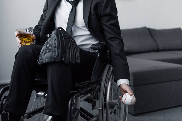 partial view of disabled man sitting in wheelchair with bag on knee while holding ball and glass of whiskey - Foto, afbeelding