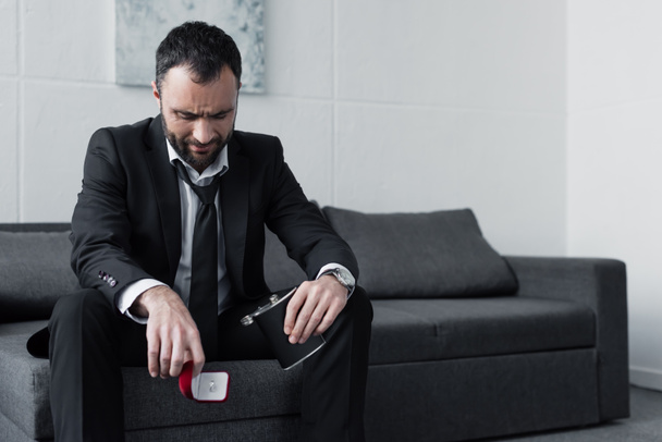 depressed bearded man sitting on sofa while holding gift box and flask  - Photo, Image