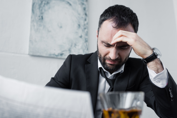 depressed businessman holding newspaper while sitting at workplace near glass of whiskey - Zdjęcie, obraz