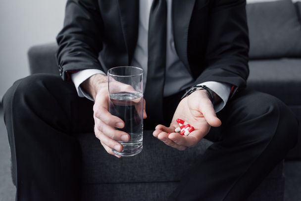 bijgesneden beeld van de mens in zwart pak die glas water en handvol pillen vasthoudt - Foto, afbeelding