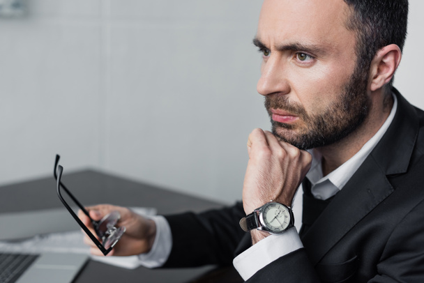 upset bearded businessman sitting at workplace, holding glasses and looking away - Zdjęcie, obraz
