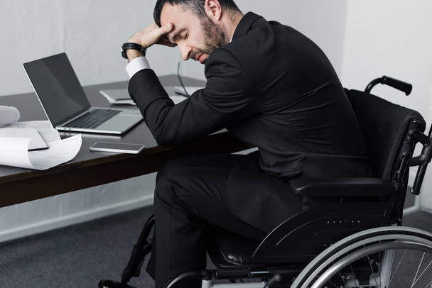депрессивный бизнесмен-инвалид в инвалидной коляске сидит на рабочем месте с ноутбуком, смартфоном и бумагами
 - Фото, изображение