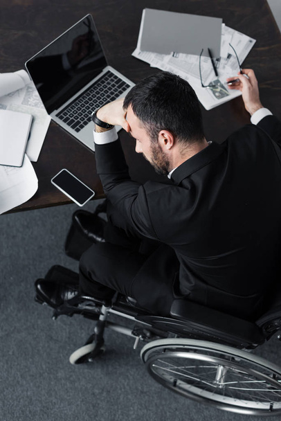 Widok z góry niepełnosprawny biznesmen siedzący w miejscu pracy w pobliżu dokumentów, laptopa i smartfona z pustym ekranem - Zdjęcie, obraz