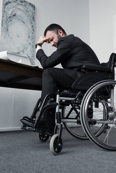 χαμηλή οπτική γωνία του καταθλιπτικός επιχειρηματίας με αναπηρία που κάθεται σε αναπηρικό καροτσάκι στο χώρο εργασίας με σκυμμένο κεφάλι - Φωτογραφία, εικόνα