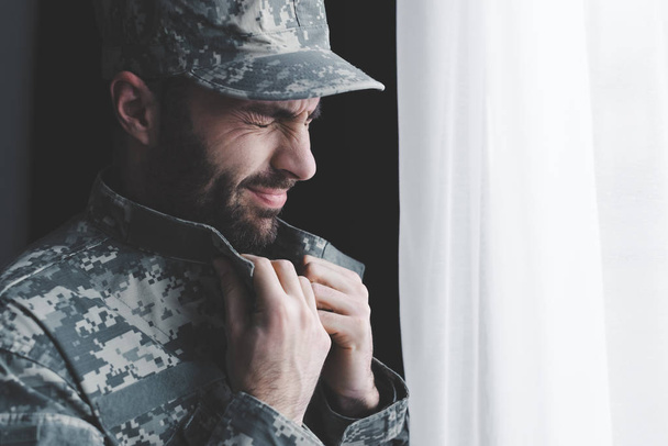 depressed bearded man in military uniform crying while standing by window - Foto, Bild