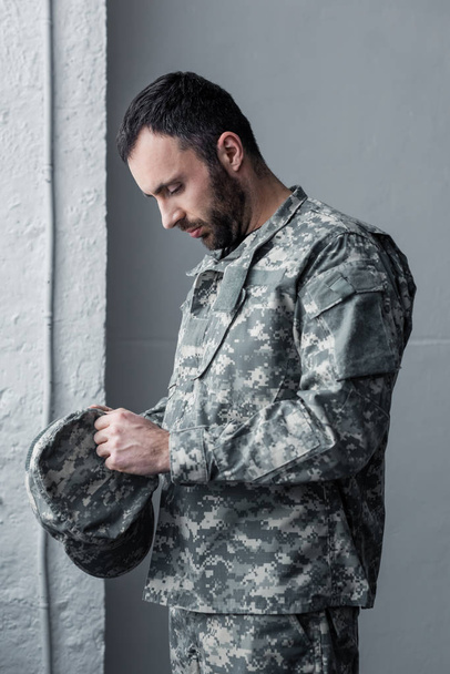 triest bebaarde man in militaire uniform staande met gebogen had en houden GLB in handen - Foto, afbeelding
