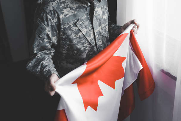 vista parcial del hombre en uniforme militar que sostiene la bandera nacional de Canadá mientras está de pie por la ventana
 - Foto, Imagen