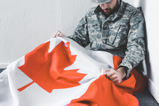 depressieve man in militair uniform zittend op de vloer in de hoek en het houden van Canada nationale vlag - Foto, afbeelding