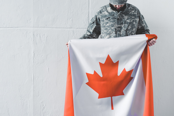 άνθρωπος με στρατιωτική ομοιόμορφη κρατώντας την εθνική σημαία του Καναδά, ενώ στέκεται κοντά σε λευκό τοίχο με κυρτό κεφάλι - Φωτογραφία, εικόνα