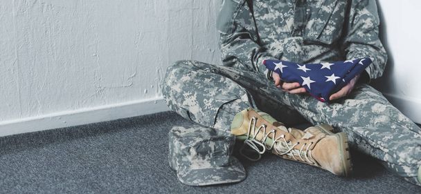bijgesneden weergave van de mens in militair uniform zittend op de vloer in de hoek en holding USA nationale vlag - Foto, afbeelding