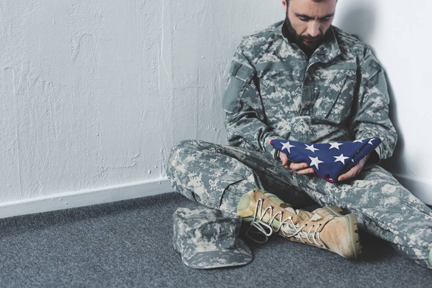 triest bebaarde man in militair uniform zittend op de vloer in de hoek en holding USA nationale vlag - Foto, afbeelding