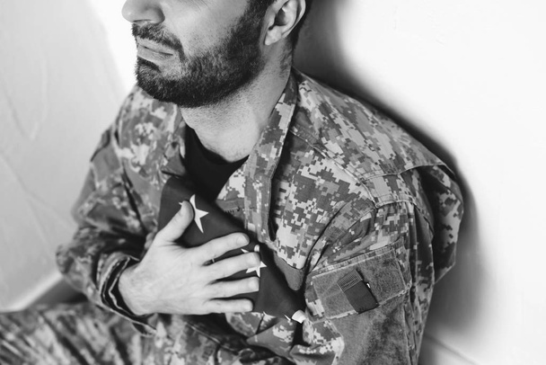 vista cortada de homem militar deprimido sentado à beira da parede branca e segurando EUA homem militar perto do coração
 - Foto, Imagem