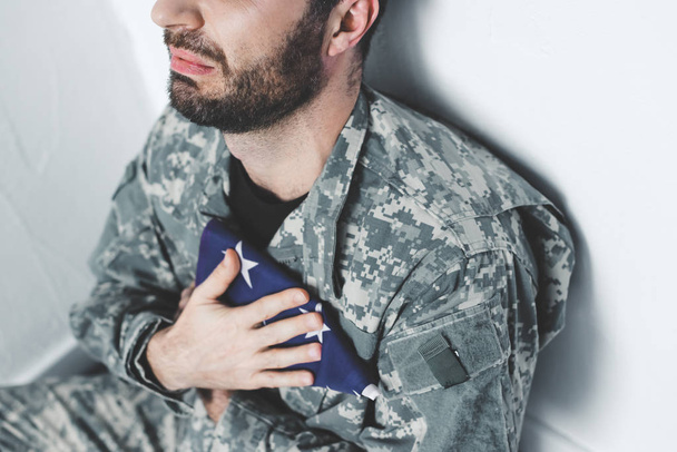 partial view of depressed military man sitting by white wall and holding usa military man near heart - Fotografie, Obrázek