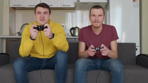 Dwóch mężczyzn grają gry wideo ze sobą. Młodzi faceci siedzą na kanapie, trzymać Gamepady w ich rękach i grać w konsoli, jeden z nich wygrywa i cieszyć się zwycięstwem, drugi przyjaciel jest zdenerwowany. materiał filmowy 4K. - Materiał filmowy, wideo
