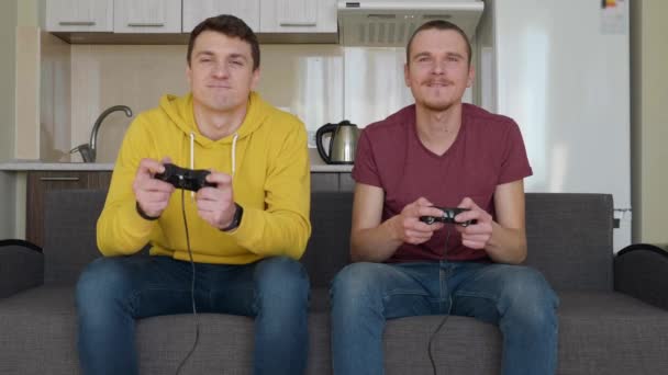 Dwóch mężczyzn grają gry wideo ze sobą. Młodzi faceci siedzą na kanapie, trzymać Gamepady w ich rękach i grać w konsole, jeden z nich wygrywa i cieszyć się zwycięstwem, drugi przyjaciel jest zdenerwowany - Materiał filmowy, wideo