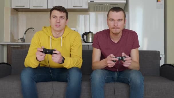 Двое мужчин с энтузиазмом играют в видеоигры. Молодые парни сидят на диване, держат в руках геймпады и играют на консоли, активно двигаются во время прохождения одного из уровней. Командный матч. 4K видео
. - Кадры, видео