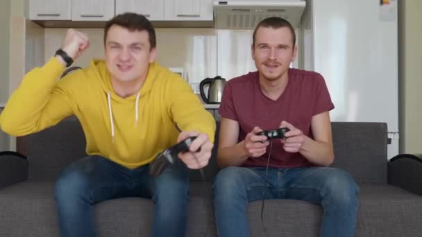 Двоє чоловіків грають у відеогру і програють. Молоді хлопці сидять на дивані, тримаючи геймпадки в руках і думають рано, що вони виграли, але вони програли врешті-решт. Друзі відпочивають. Кухня на фоні. Знімок 4K
. - Кадри, відео
