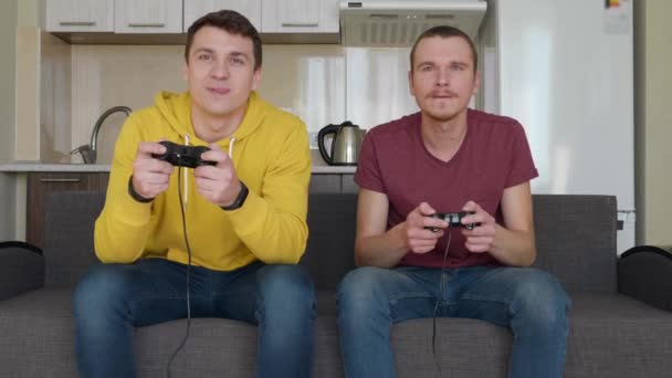 Two men are playing video game and win. Young guys are sitting on the couch, hold gamepads in their hands, play console, enjoy the victory, one of friends rises from the sofa and waves his hands. Team match. 4K footage. - Footage, Video