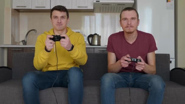 Két férfi lelkesen játszanak videójáték. Fiatal fickók van ülő-on dívány, ragaszkodik Gamepad-ban-uk kezek és játék a vigasztal, aktívan mozog közben a áthaladás-ból egy-ból szintek. Csapatmeccs. 4k felvételek. - Felvétel, videó