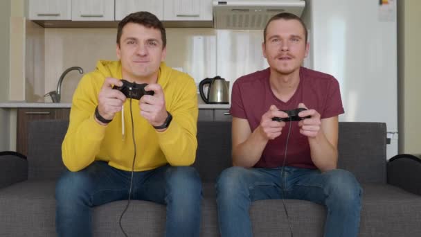 Двое мужчин с энтузиазмом играют в видеоигры. Молодые парни сидят на диване, держат в руках геймпады и играют на консоли, активно двигаются во время прохождения одного из уровней. Командный матч. 4K видео
. - Кадры, видео