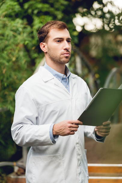 serious scientist in white coat holding folder in green orangery - Фото, изображение