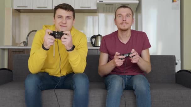 Двое мужчин играют в видеоигры и проигрывают. Молодые парни сидят на диване, держат в руках геймпады и играют на консоли, обсуждают свои ошибки во время прохождения одного из уровней. Командный матч. 4K видео
. - Кадры, видео