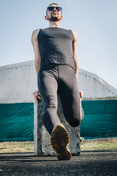 A slender young man in black clothes and sunglasses is exercising outdoors. fitness athlete on the sports field. training. vertical. warm up stretching legs. body preparation for the summer. vertical - Foto, immagini